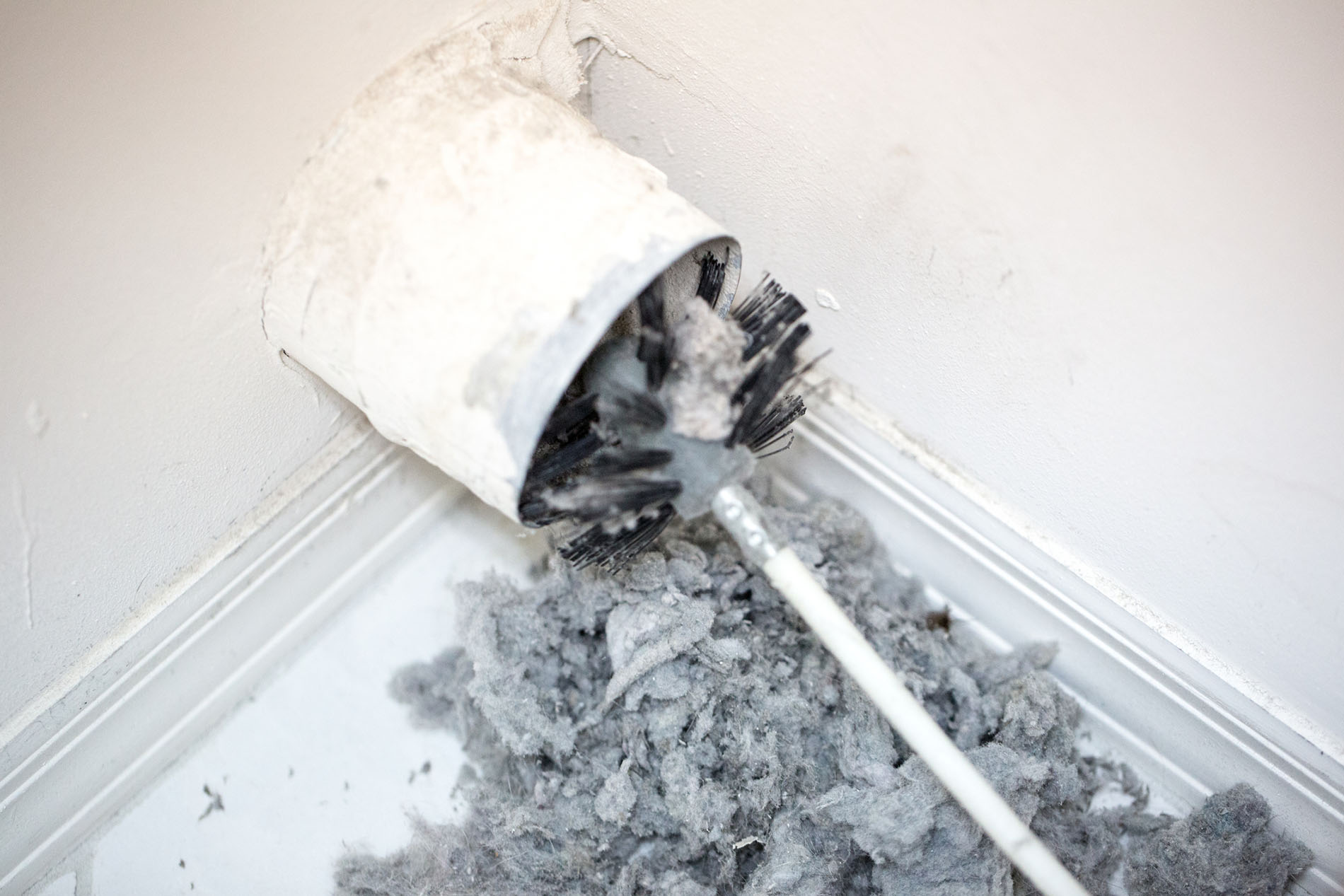 Dryer vent in a home being cleaned out with a round brush. There is a large pile of lint that has been removed from the vent on a tiled floor.
